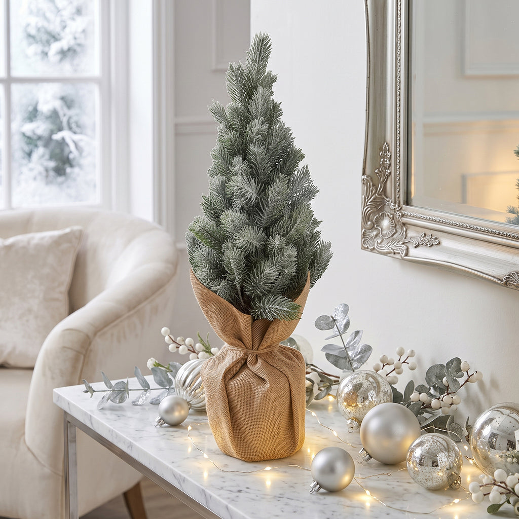 Oszroniona choinka w jucie ustawiona na marmurowym blacie konsoli, w otoczeniu srebrnych bombek, lampek i białych dodatków dekoracyjnych.